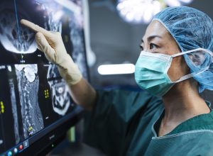 Female surgeon in scrubs and mask pointing at MRI images on a monitor during surgery preparation.