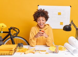 Happy dark skinned female student with curly Afro hair does hometask makes report draws sketches wears sweatshirt pose in coworking space against yellow background blueprint hanging on wall.