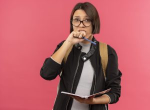 anxious young student girl wearing glasses and back bag holding note pad and pen keeping hand near mouth isolated on pink background with copy space