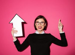 Portrait of a happy pretty businesswoman holding paper arrow sign up and pointing finger up isolated over pink background