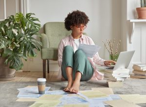 Serious black lady with Afro haircut, makes research work, checks data on laptop computer, holds documents, poses at floor in fashionable clothes, surrounded with pile of books. Student studies indoor
