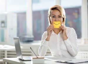 Young office worker in white shirt holding yellow notepaper with drawn smile by her mouth