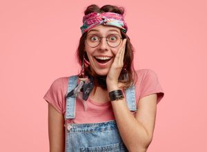 Studio shot of stylish hippy female has broad smile, keeps hand on cheek, eyes popped out, wears round spectacles, can not believe in success, wears denim overalls, models against pink background